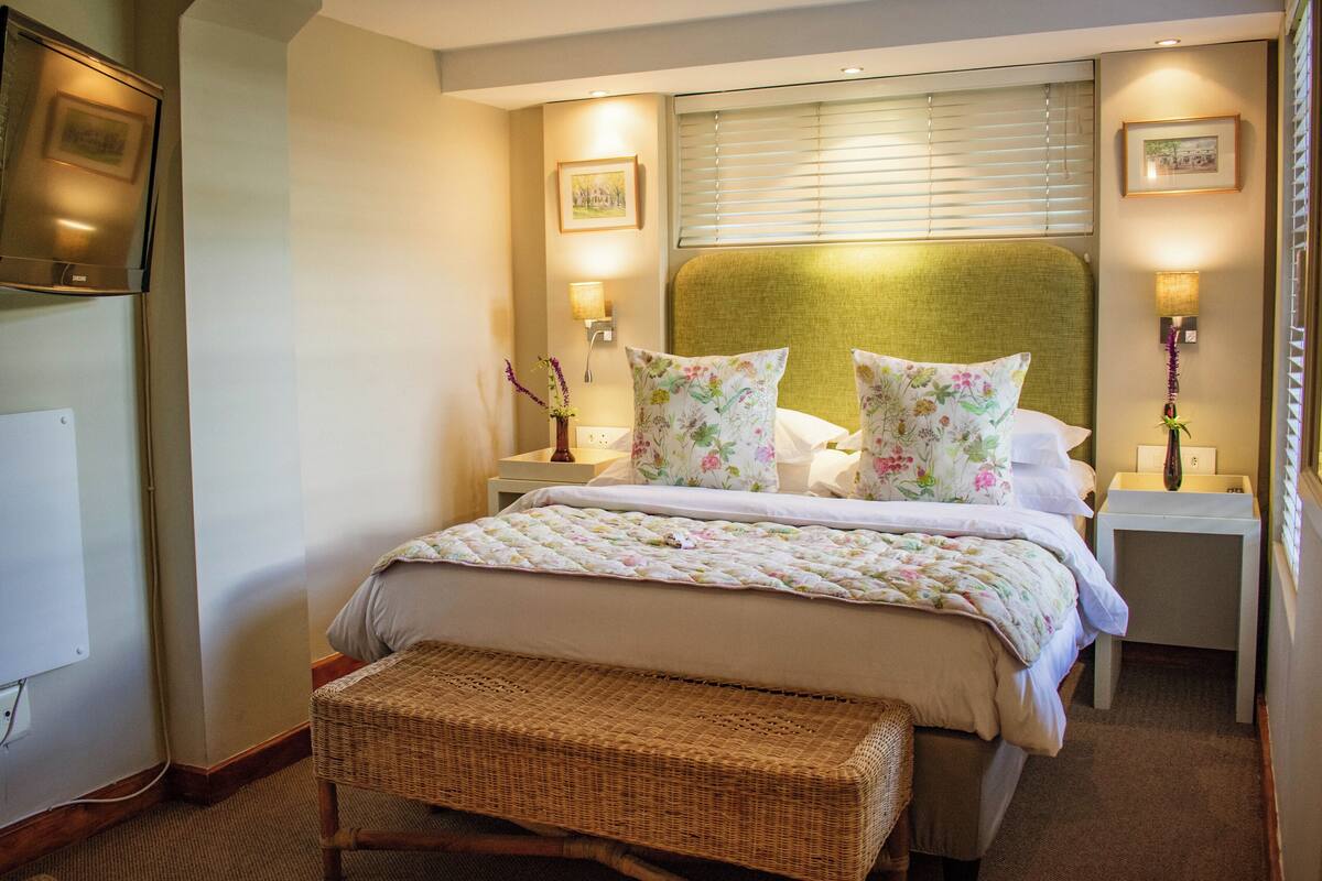 A hotel room with a large bed, a bedside table, a nightstand, a television, and a window with blinds.