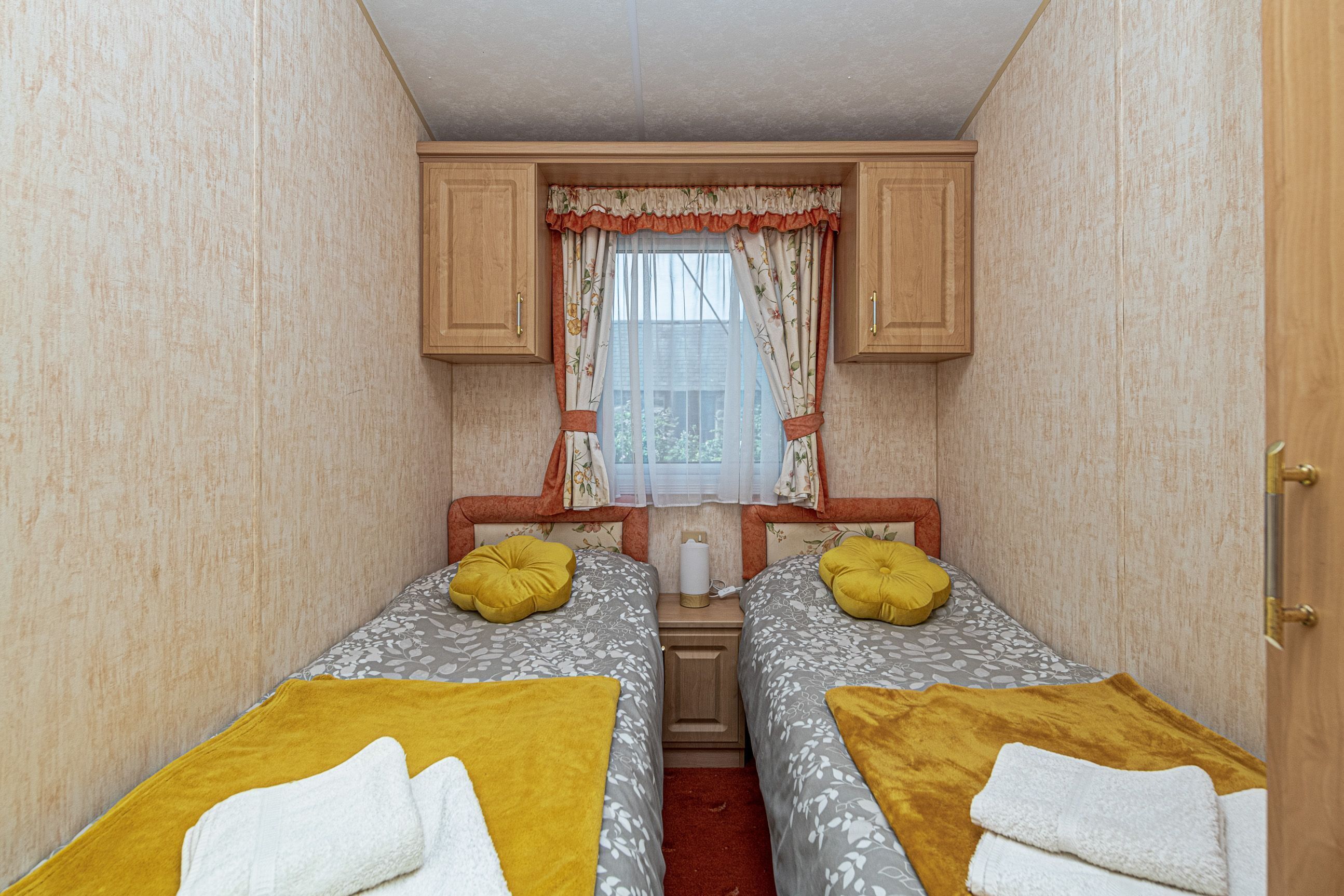 Une chambre de taille modeste, équipée de deux lits, chacun agrémenté d’un oreiller jaune et d’une serviette blanche. Une fenêtre, ornée de rideaux à motifs floraux, et une petite table se trouvent entre les deux lits.