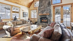 A cozy living room with a stone fireplace, leather sofas, and large windows with snow views. - Silverado Lodge at Palisades - 4BR w/ Private Hot Tub, Ski Shuttle & Pet-Friendly! (Olympic Valley)