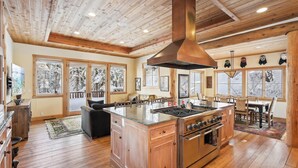A spacious kitchen with wooden flooring, a central island with a stove, and large windows with snowy views. - Silverado Lodge at Palisades - 4BR w/ Private Hot Tub, Ski Shuttle & Pet-Friendly! (Olympic Valley)
