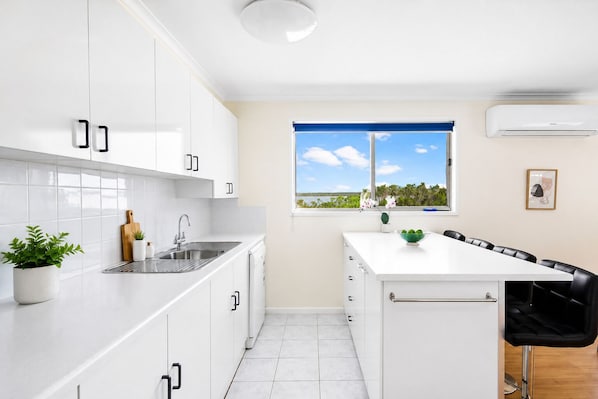 A modern kitchen with white cabinets, a stainless steel sink, and a window with a view of the outdoors. - Carlenes Getaway | Lake Views, Beach & Games Hub (Loch Sport)