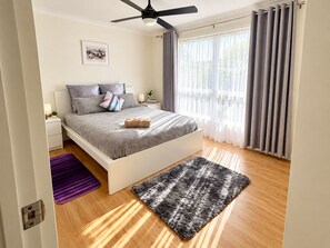 Quarto moderno com cama, ventilador de teto, janela com cortinas e uma pequena mesa de cabeceira.