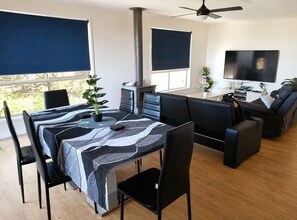 A modern living room with a dining area, a fireplace, a flat-screen TV, and a ceiling fan. - Carlenes Getaway | Lake Views, Beach & Games Hub (Loch Sport)