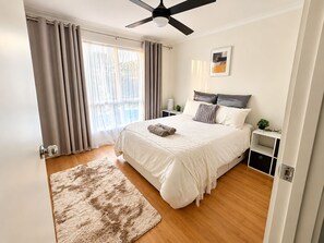 A bedroom with a bed, a ceiling fan, a bedside table with a plant, and a window with curtains. - Carlenes Getaway | Lake Views, Beach & Games Hub (Loch Sport)