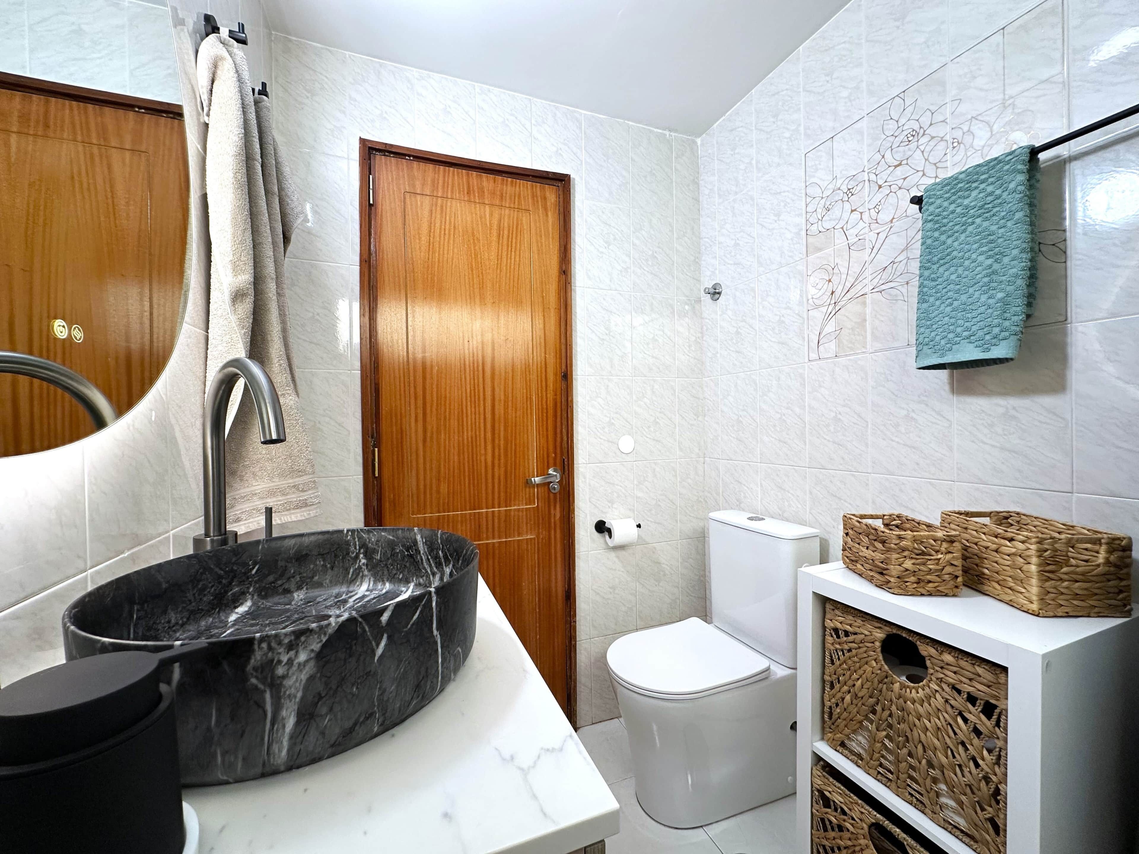 Un baño con lavamanos de mármol, puerta de madera, inodoro blanco y un perchero con una toalla azul.