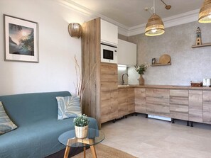 Een moderne woonkamer met een blauwe bank, een glazen salontafel en een keuken met houten kastjes en een inbouwmagnetron.