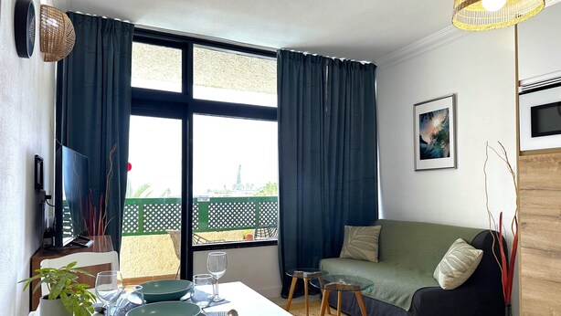 Un salon moderne comprenant un coin repas, de grandes fenêtres avec des rideaux, un canapé, un téléviseur et une plante.