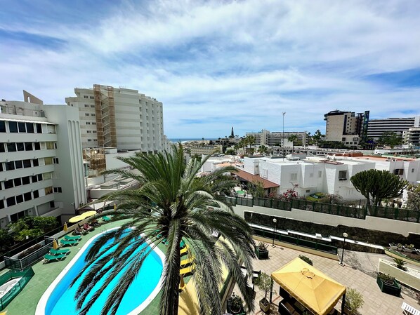 Un área de piscina en la azotea con palmeras, cielo despejado y edificios circundantes.