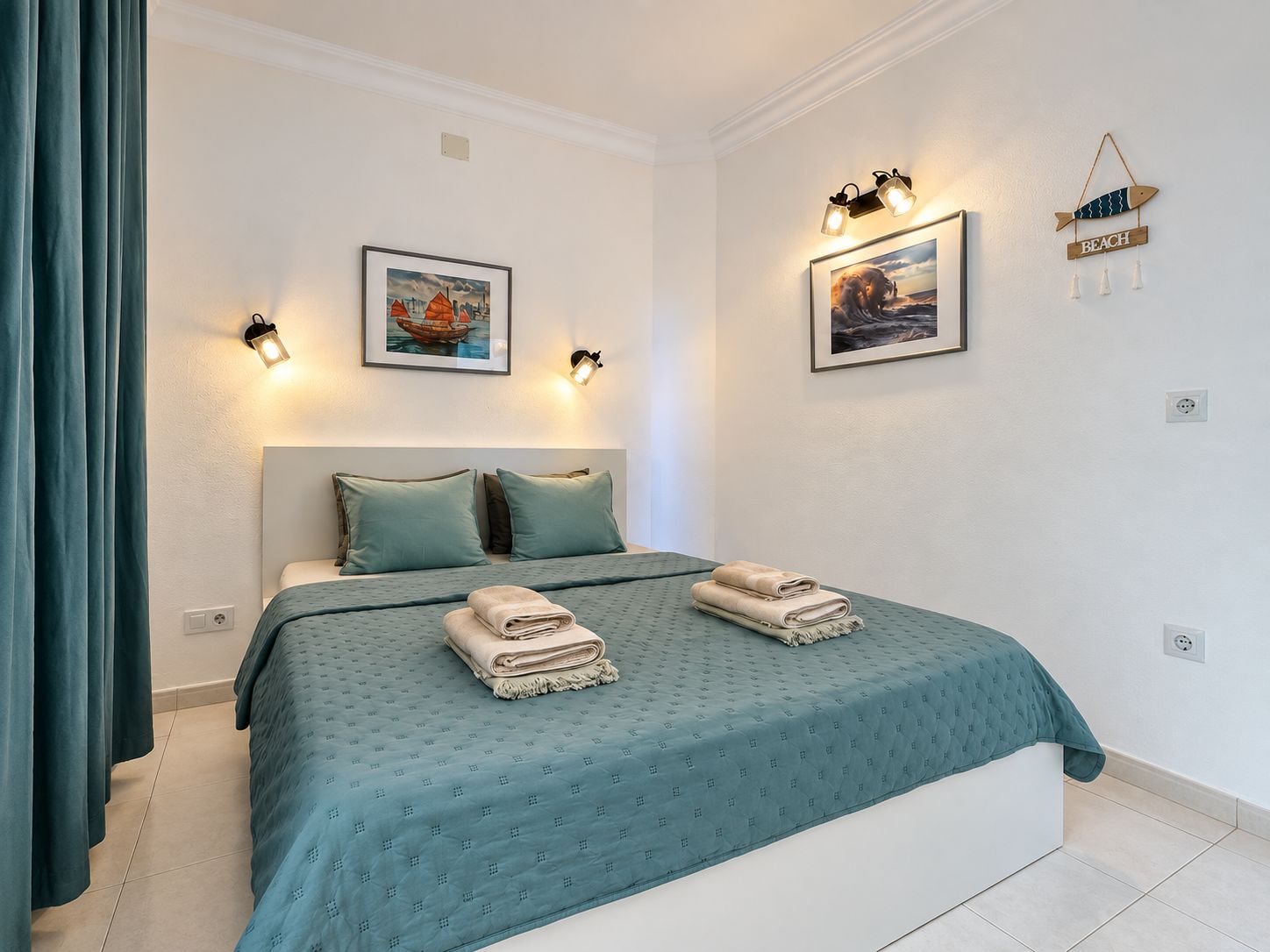 Un dormitorio con cama, dos almohadas, colcha color turquesa, dos toallas dobladas, luces empotradas en la pared y dos cuadros enmarcados.