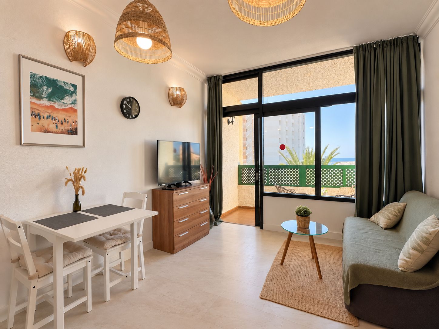A modern living room with a sofa, a dining table with chairs, a TV, and a large window with a view of the beach.