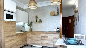 Une cuisine moderne avec un îlot central en bois, des armoires blanches et un micro-ondes encastré. On y trouve des suspensions en osier, une petite étagère avec des objets décoratifs, ainsi qu’une table à manger dressée avec de l’assiette et des verres bleus.