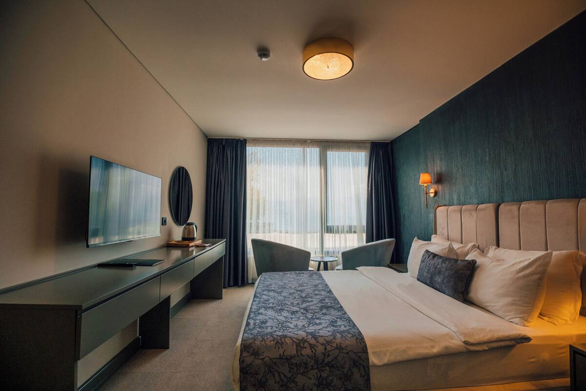 A hotel room with a bed, bedside table, lamp, and a large window with curtains.