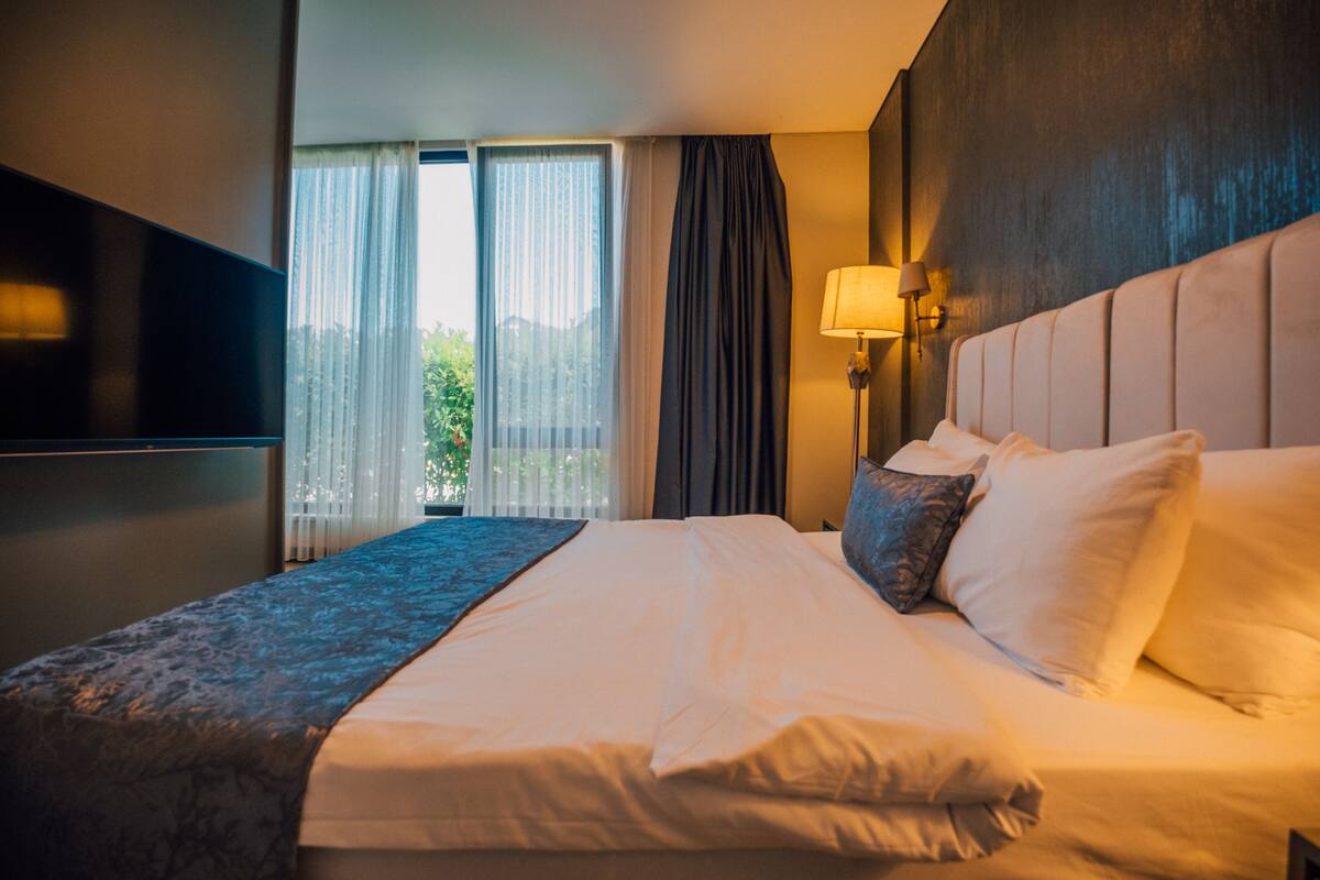 A hotel room with a large bed, a flat-screen TV mounted on the wall, a bedside lamp, and a view of greenery outside.