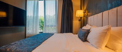 A hotel room with a large bed, a flat-screen TV mounted on the wall, a bedside lamp, and a view of greenery outside.