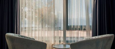 A room with two armchairs and a small table, a large window with sheer curtains, and a view of an outdoor area.