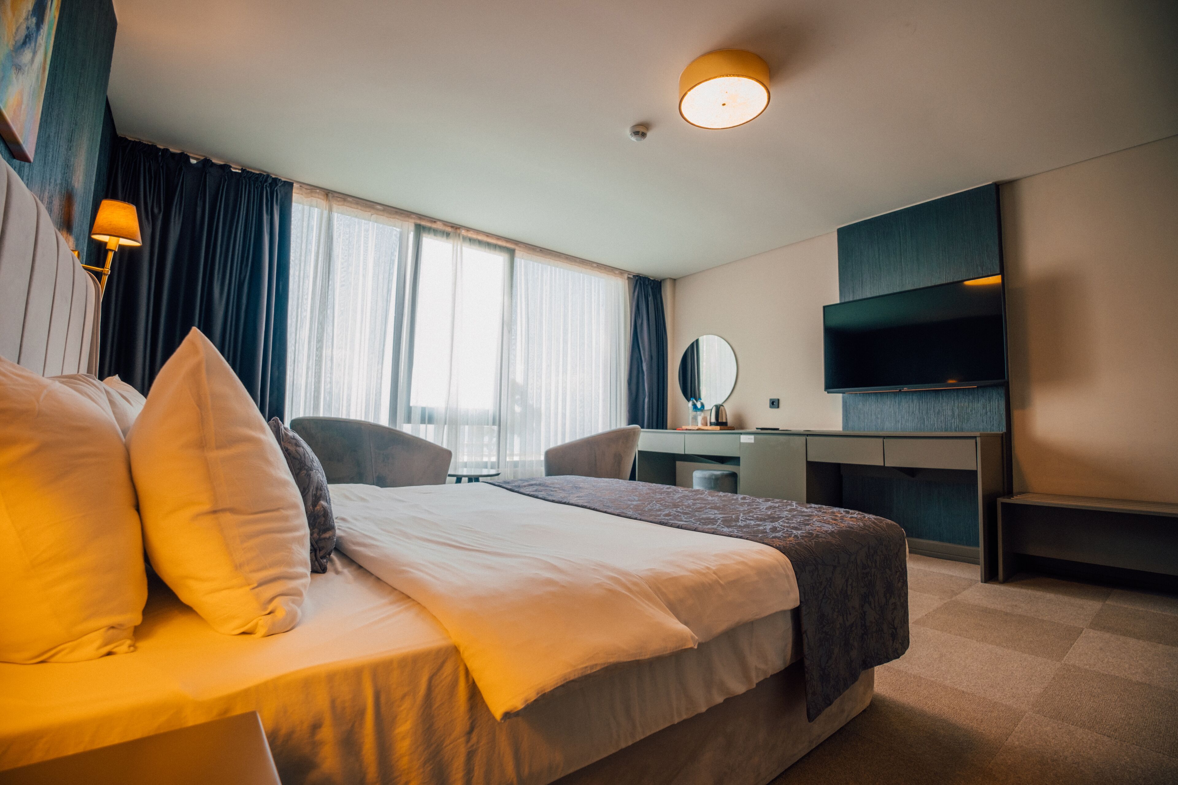 A hotel room with a large bed, a desk, a chair, a TV, and a window with curtains.