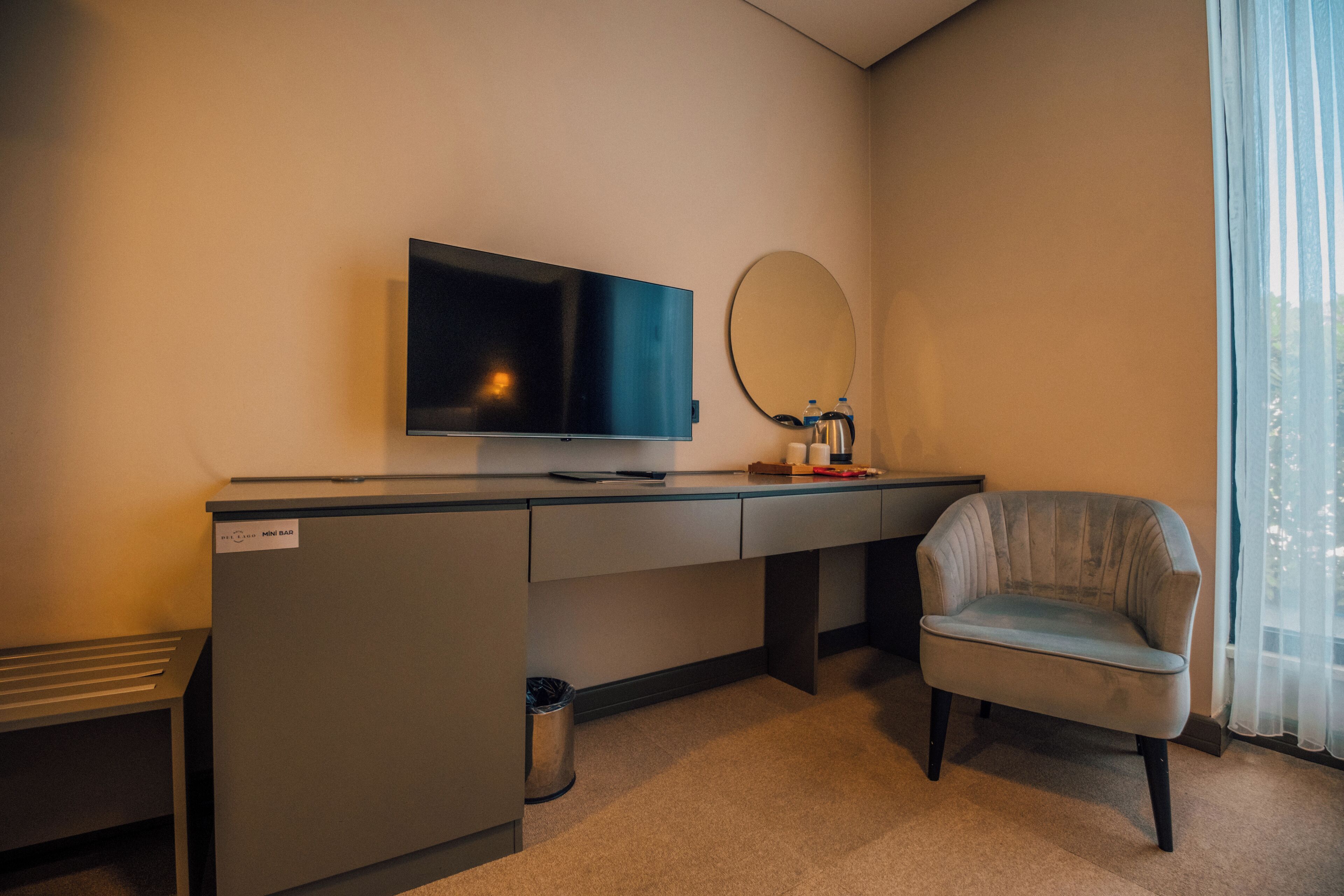 Habitación de hotel con televisor de pantalla plana, espejo redondo, sillón gris y minibar.