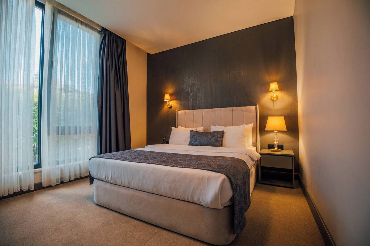 A hotel room with a large bed, two bedside lamps, a nightstand, and a view of greenery through sheer curtains.