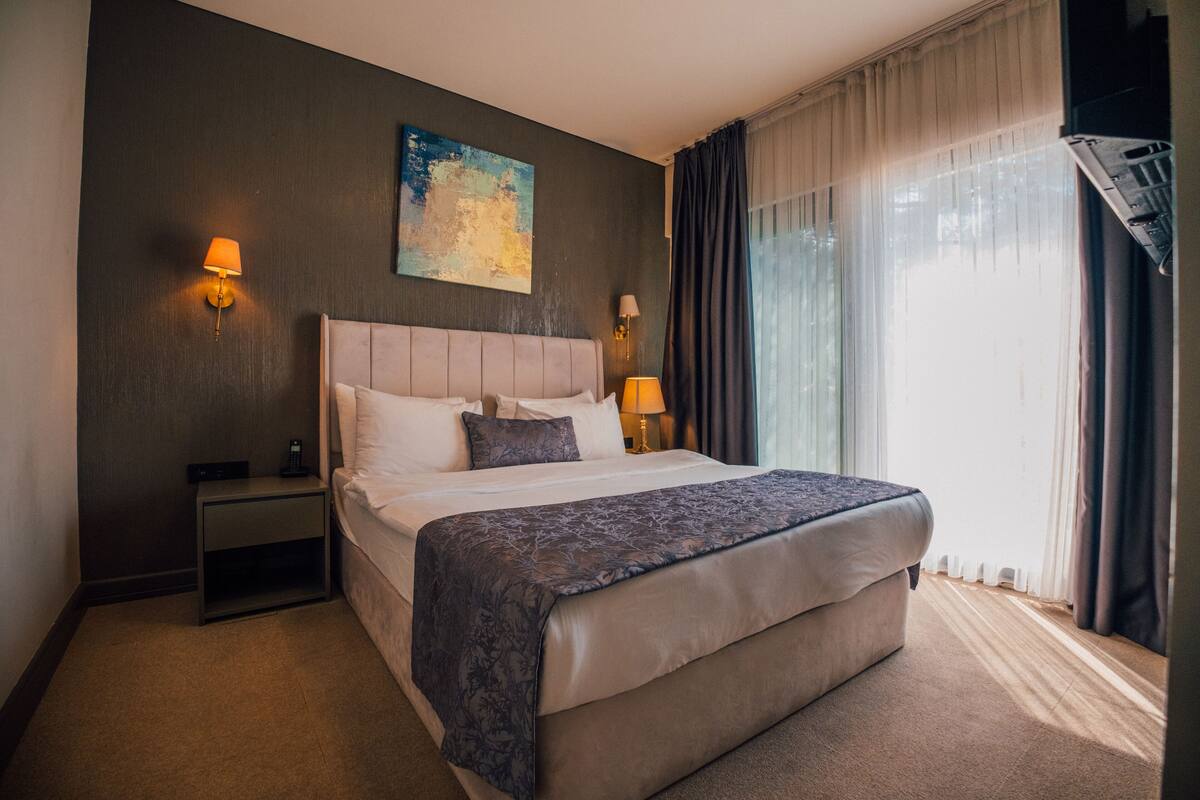 A hotel room with a large bed, two bedside tables with lamps, a painting on the wall, and a view of the outdoors through sheer curtains.