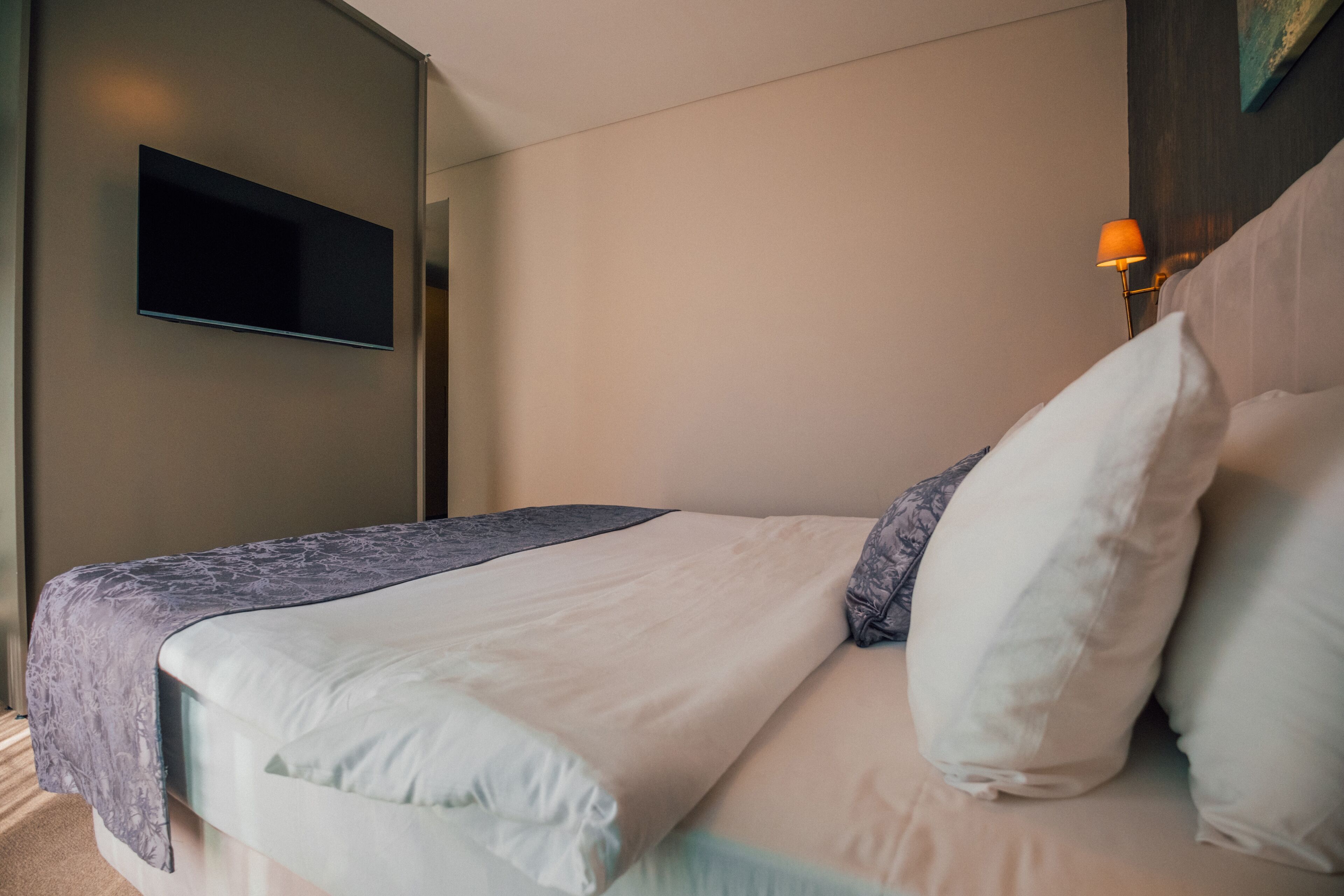 A neatly made bed with a white and light blue bedspread, a flat-screen TV mounted on the wall, and a bedside table with a lamp.