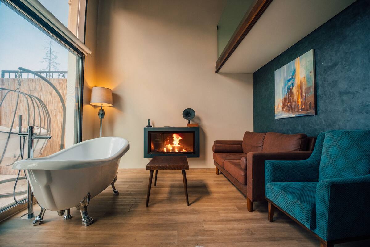 A modern living room with a freestanding bathtub, a fireplace, a sofa, and a teal armchair.