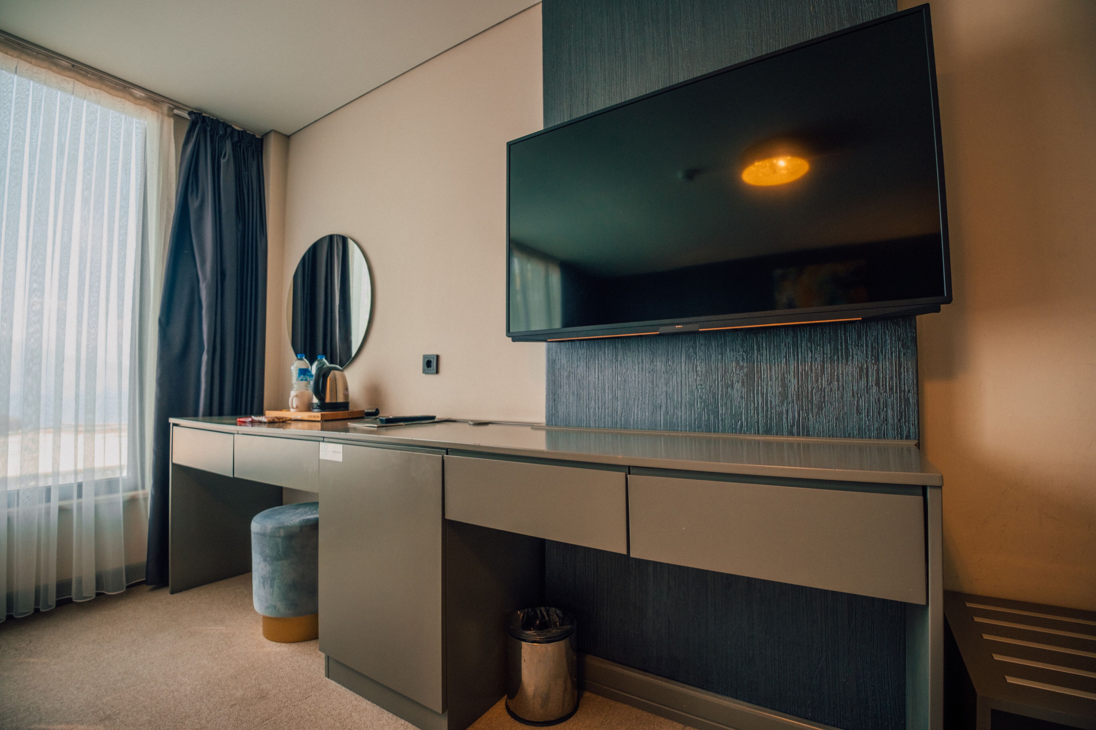 Ein modernes Hotelzimmer mit großem Schreibtisch, Flachbildfernseher an der Wand, rundem Spiegel und kleiner Mülleimer.