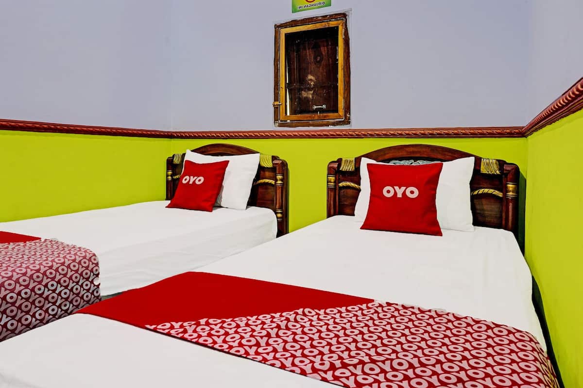 Une chambre d’hôtel avec deux lits, chacun doté d’une tête de lit en bois, de draps blancs et de couvertures à motifs rouges et blancs. Une petite fenêtre avec un cadre en bois est visible sur le mur.