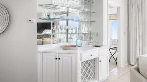 A modern kitchen with white cabinetry, a glass wine rack, and a built-in sink. - Epic 4BR/4BA Gulf Front | Free Beach Chair Service (Navarre)