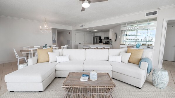 A spacious living room with a large white sectional sofa, a wicker coffee table, and a dining area with white chairs. - Epic 4BR/4BA Gulf Front | Free Beach Chair Service (Navarre)