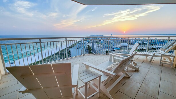 A balcony with white chairs and a table, overlooking a beach and cityscape during sunset. - Epic 4BR/4BA Gulf Front | Free Beach Chair Service (Navarre)