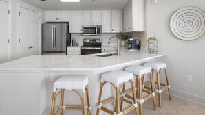 A modern kitchen with white cabinetry, stainless steel appliances, a marble countertop, and wicker bar stools. - Epic 4BR/4BA Gulf Front | Free Beach Chair Service (Navarre)