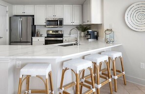Una cocina moderna con armarios blancos, electrodomésticos de acero inoxidable, una encimera de mármol y tres taburetes de mimbre.