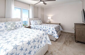 A hotel room with two beds, a ceiling fan, a nightstand with a lamp, and a view of the ocean.