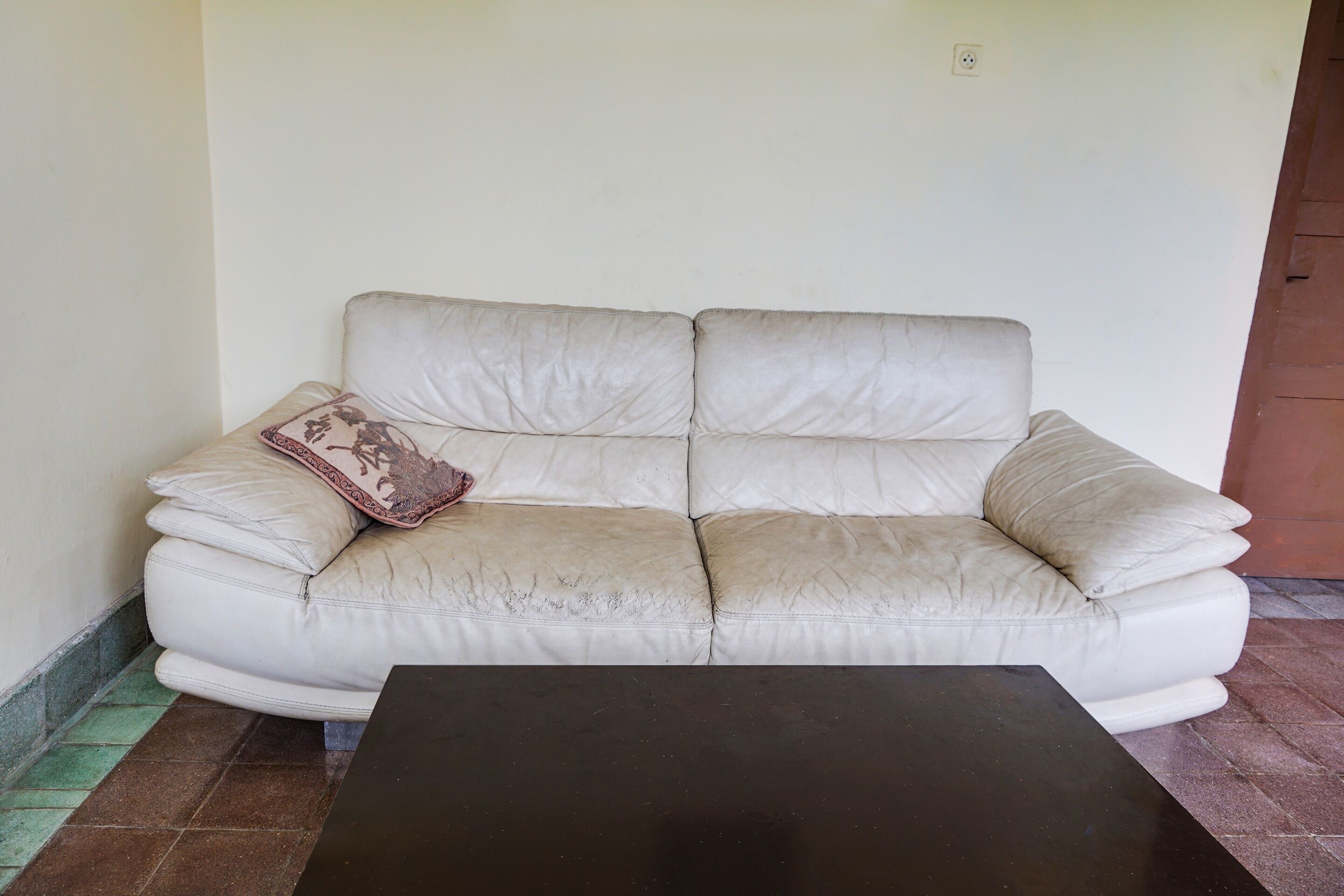 A white leather sofa with a patterned pillow against a plain wall, a black table in front, and a tiled floor.