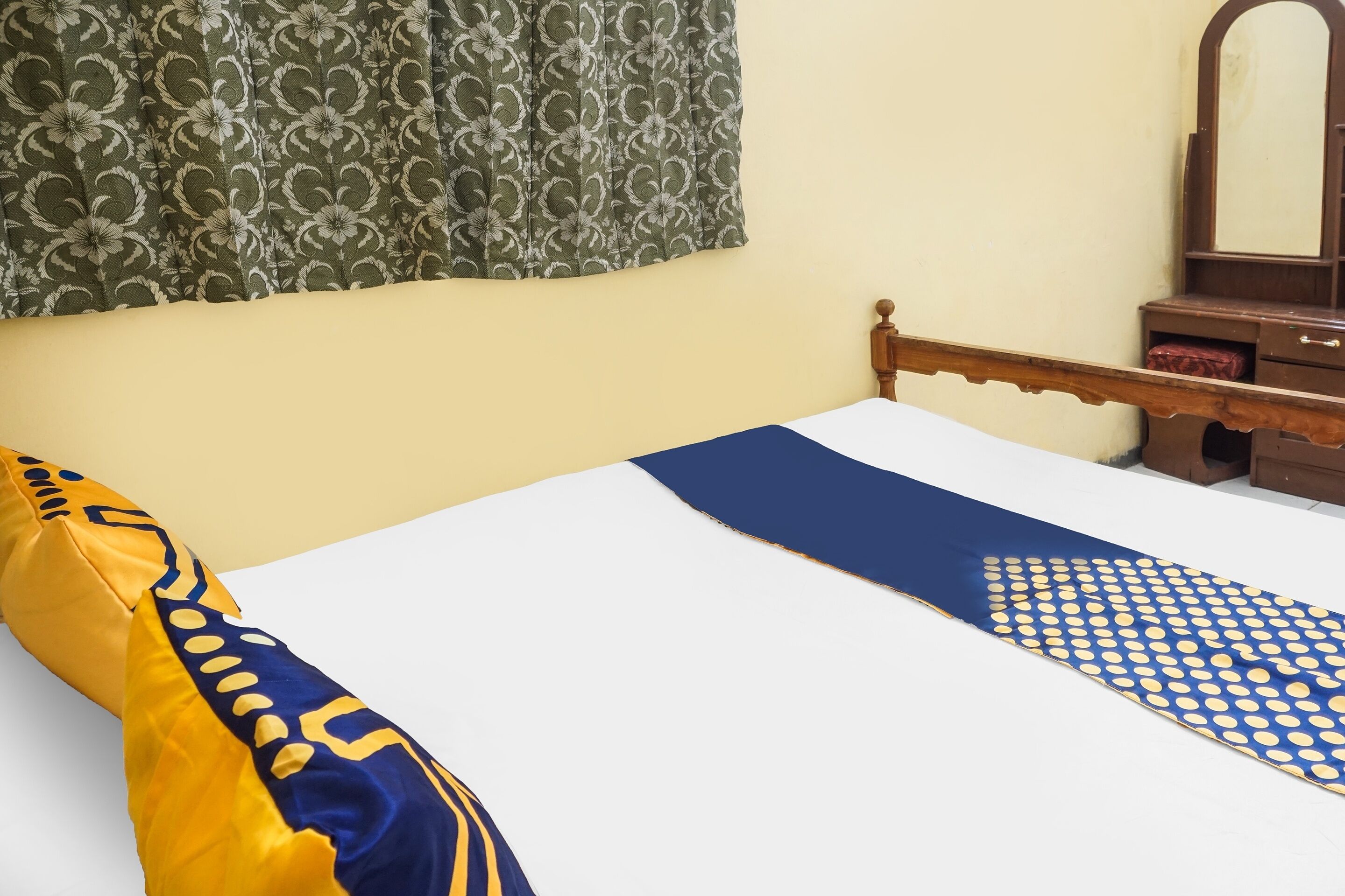 Una cama con colcha blanca y azul, almohada amarilla y azul, y cortina estampada.