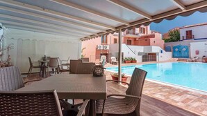 A covered outdoor area with a pool, wicker furniture, and a view of nearby buildings.