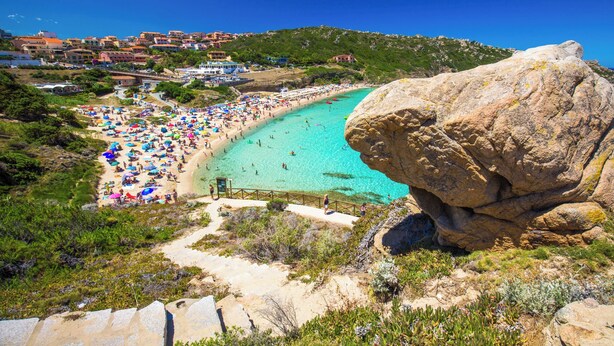 A coastal beach with a large rock formation, clear turquoise water, and a path leading to the beach.
