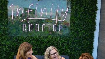 Three people seated on outdoor furniture with a hedge wall and a neon sign reading 'Infinity Rooftop' behind them.