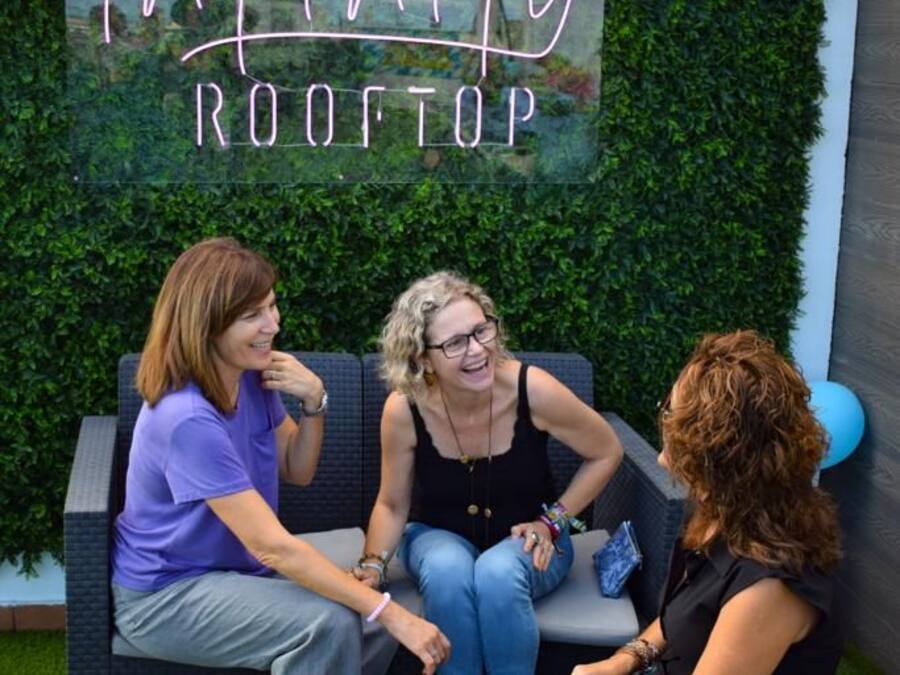 Three people seated on outdoor furniture with a hedge wall and a neon sign reading 'Infinity Rooftop' behind them.
