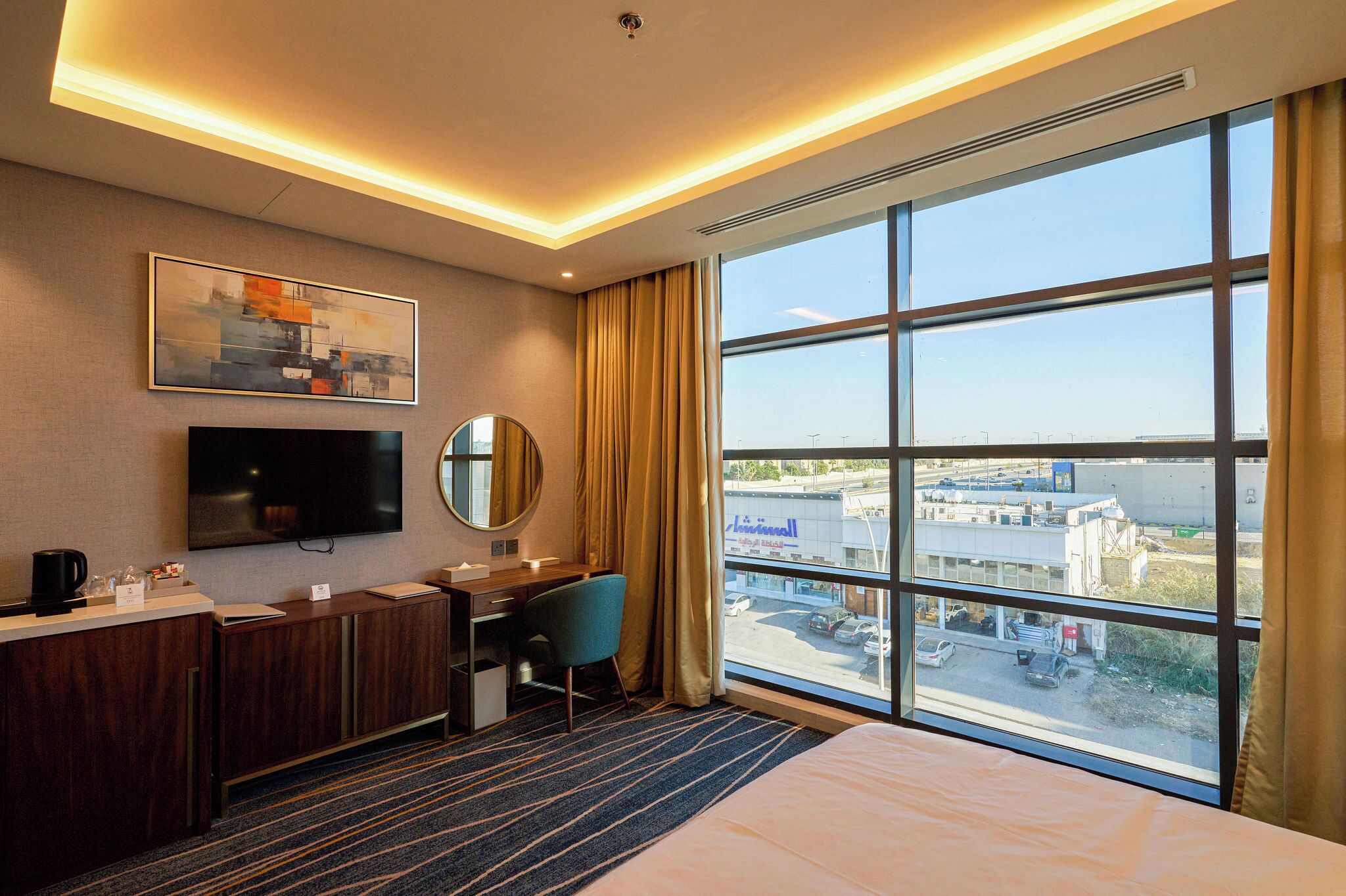 A hotel room with a large window, a desk, a chair, a bed, a TV, and a painting on the wall.