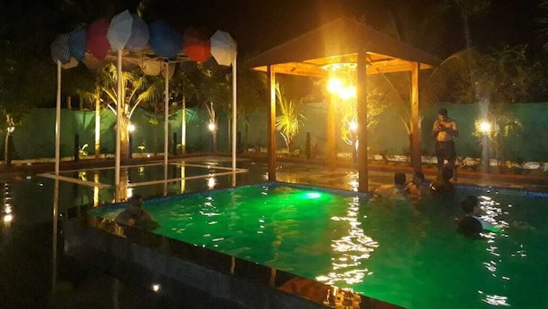 A nighttime swimming pool area with a covered pavilion, a water fountain, and a person swimming. - Sealand Kihim (Alibag)