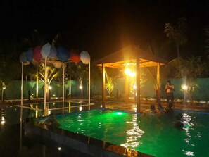 Un espace piscine nocturne doté d’un pavillon couvert, d’une fontaine et d’une personne qui nage.