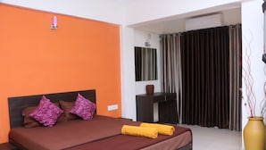 A hotel room with an orange wall, a brown bed with purple and yellow pillows, a desk, a mirror, and a large vase with branches. - Sealand Kihim (Alibag)