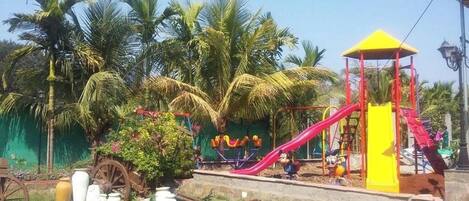 Un parque infantil con un módulo de juegos amarillo, un tobogán rojo y palmeras al fondo.