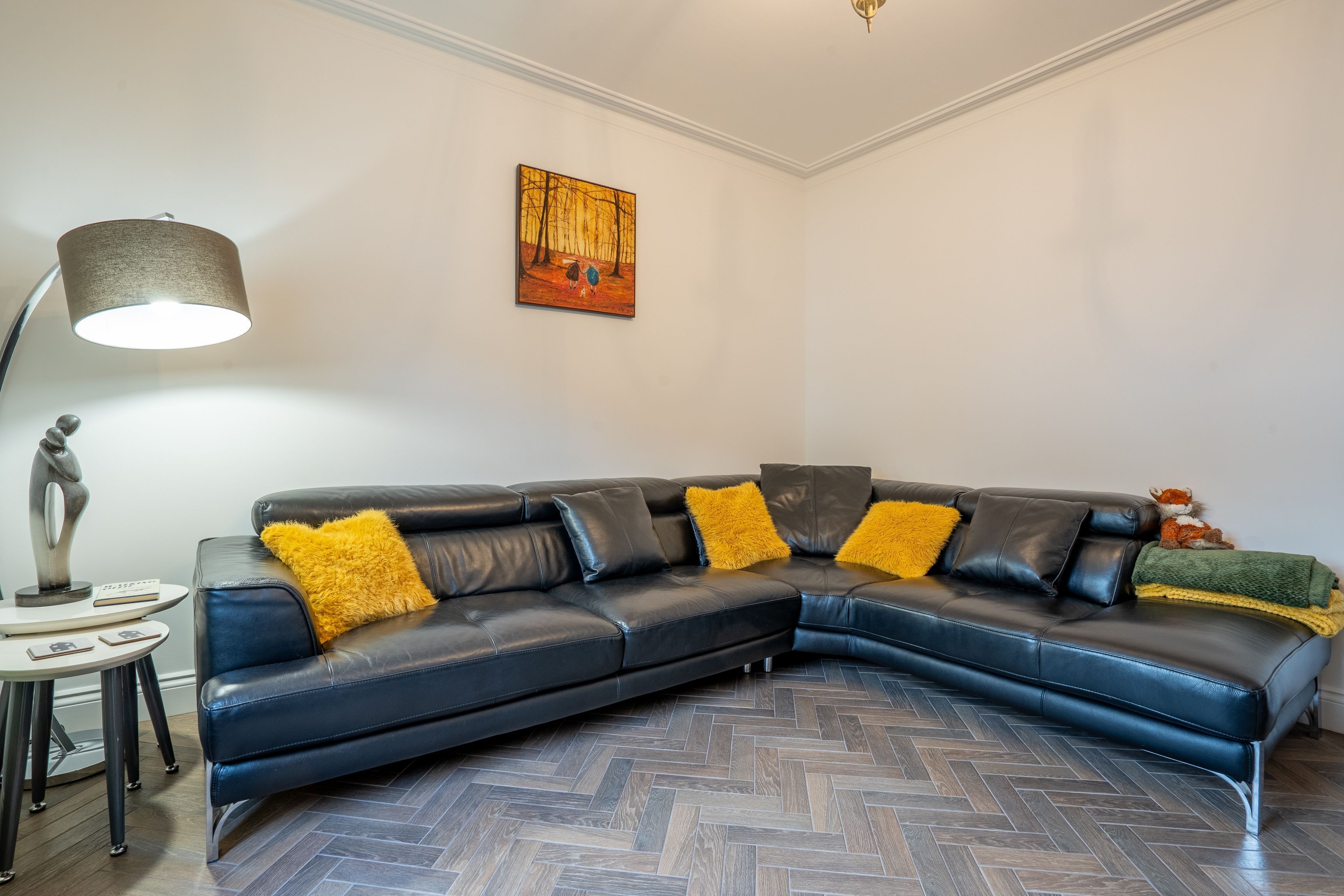 A modern living room with a black leather sectional sofa, a round table with a lamp, and a herringbone-patterned floor.