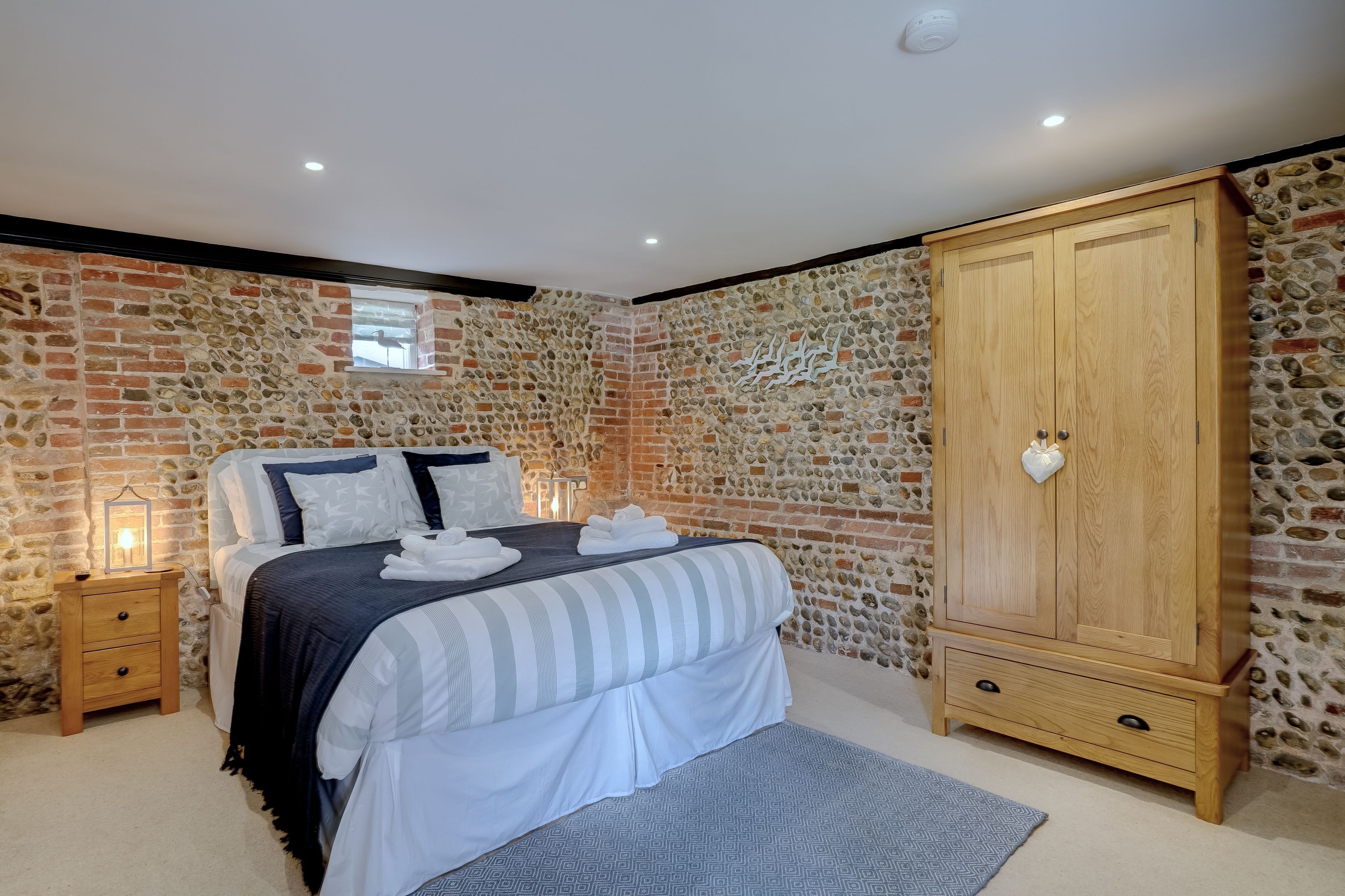 Un dormitorio con pared de piedra, un armario de madera, una cama con ropa de cama blanca y una mesita de noche con lámpara.