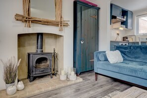 Een woonkamer met een stenen openhaard, een blauwe bank en een keuken met kasten en een raam.