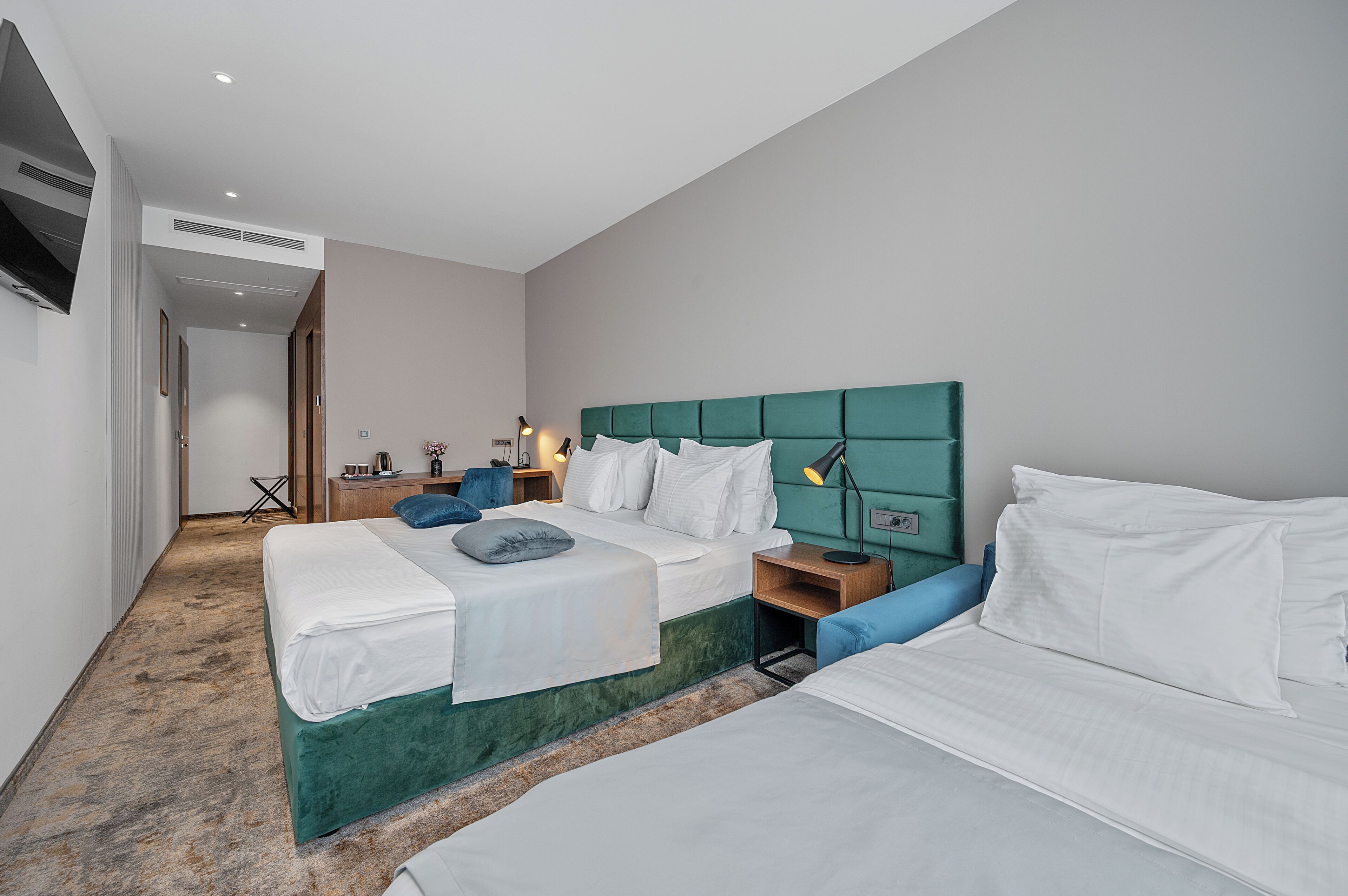 Habitación de hotel con dos camas, cabecero verde, ropa de cama blanca, un escritorio pequeño con una lámpara y un televisor montado en la pared.