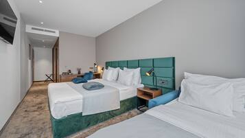 Habitación de hotel con dos camas, cabecero verde, ropa de cama blanca, un escritorio pequeño con una lámpara y un televisor montado en la pared.