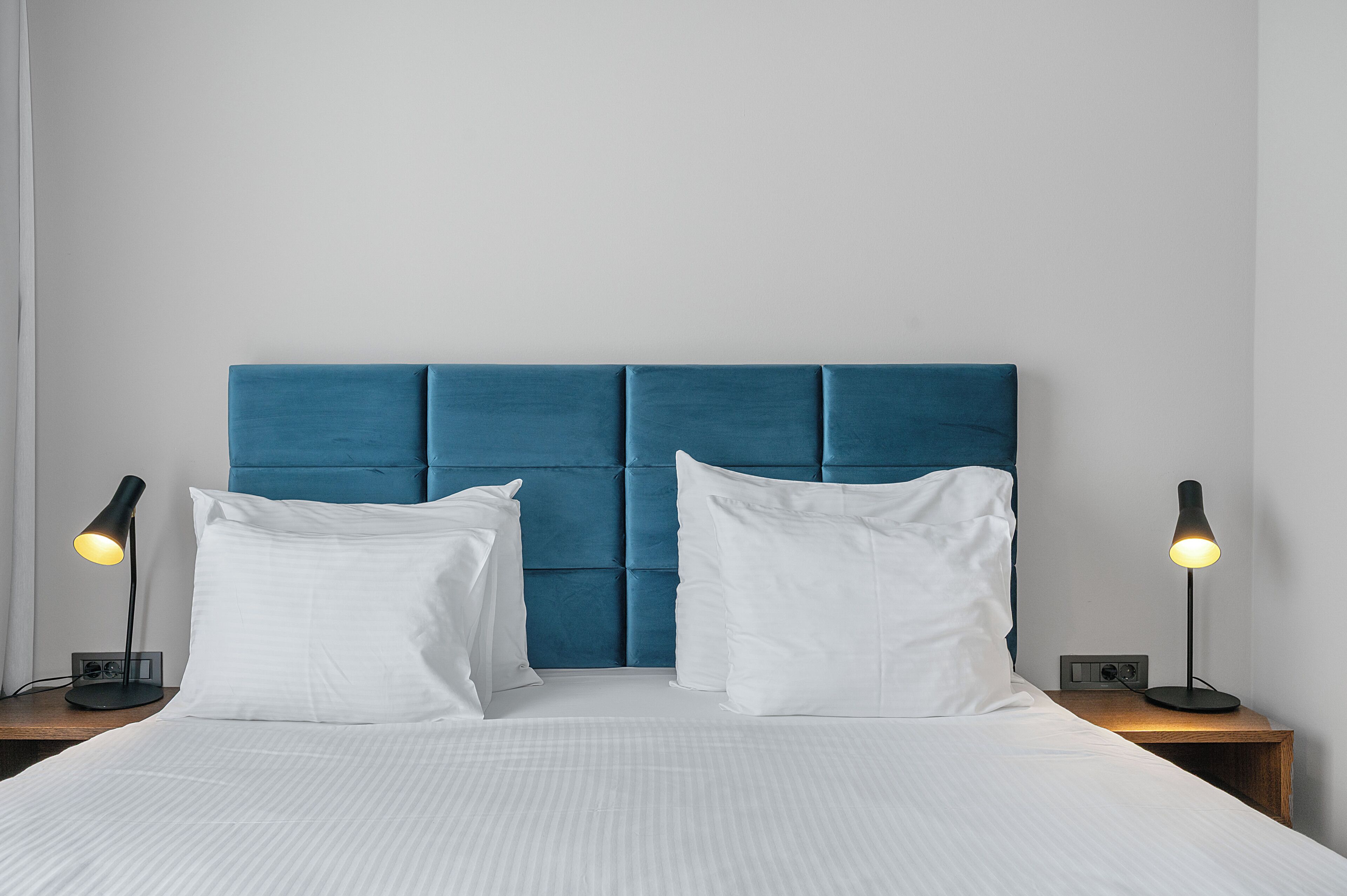 Una cama bien tendida con sábanas blancas, un cabecero tapizado en azul y dos lámparas de noche sobre mesitas de madera.